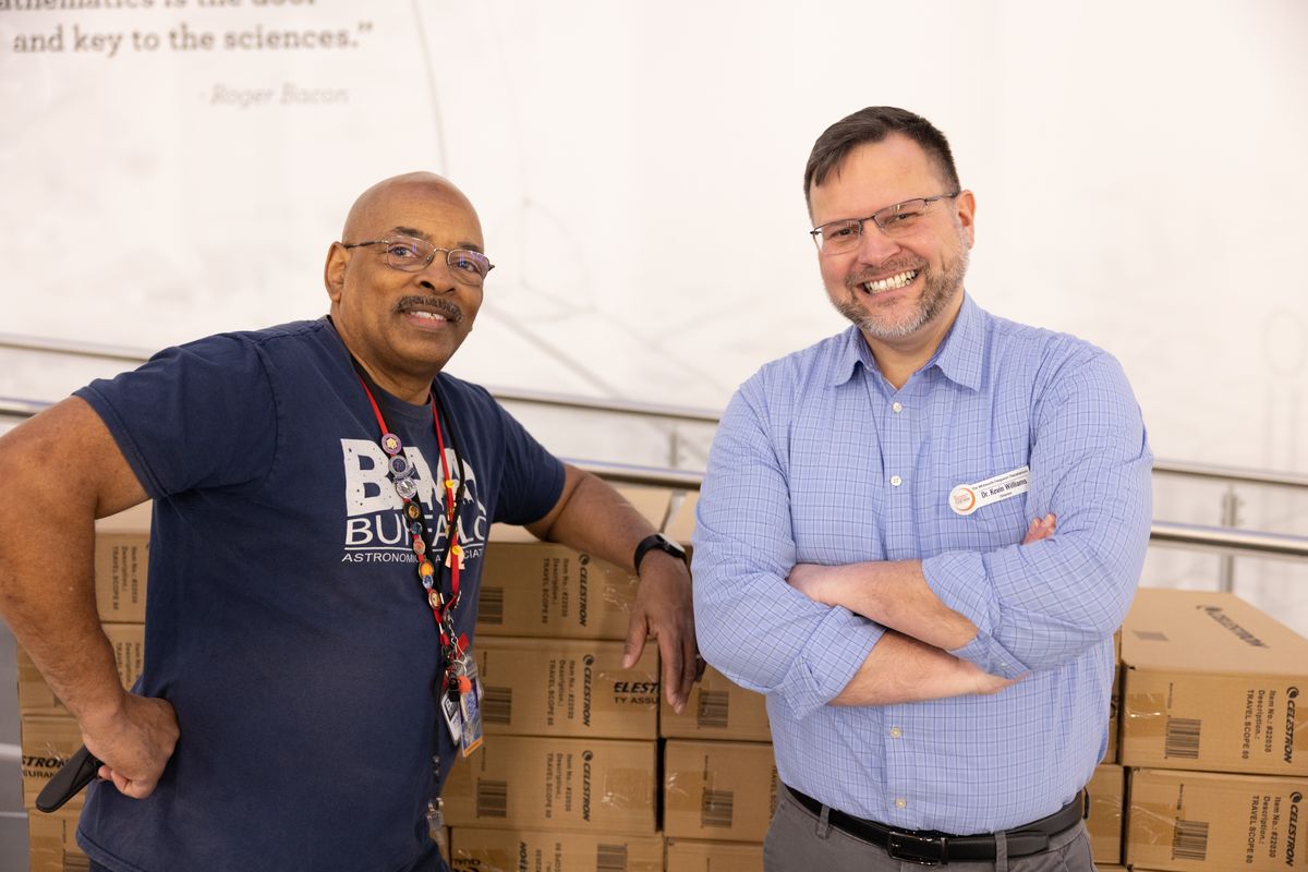 Michael Humphrey, in a Buffalo Astronomical Association shirt, and Dr. Kevin Williams, wearing his name tag, stand in front of stacked Celestron telescope boxes