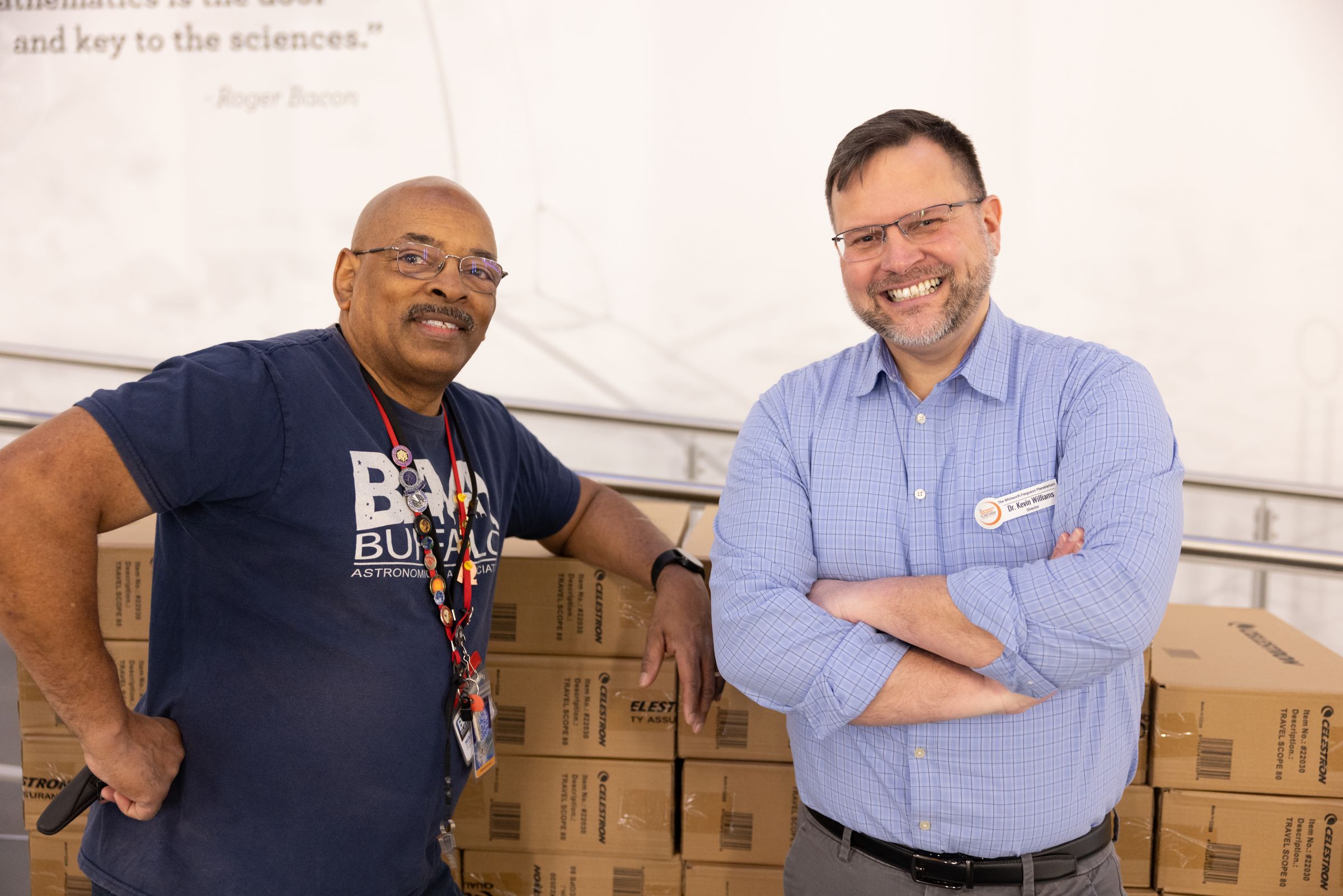 Michael Humphrey, in a Buffalo Astronomical Association shirt, and Dr. Kevin Williams, wearing his name tag, stand in front of stacked Celestron telescope boxes