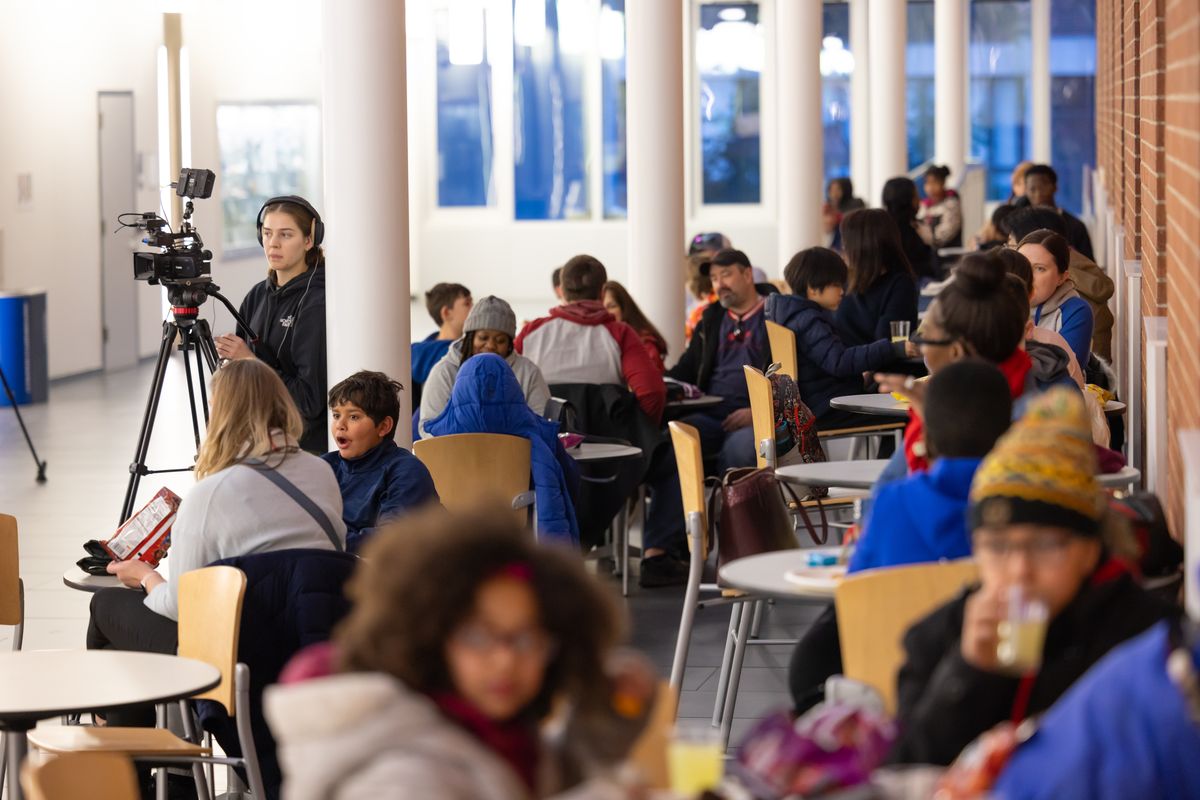 Families fill the atrium tables during the pizza party while a videographer in headphones stands by a camera on a tripod