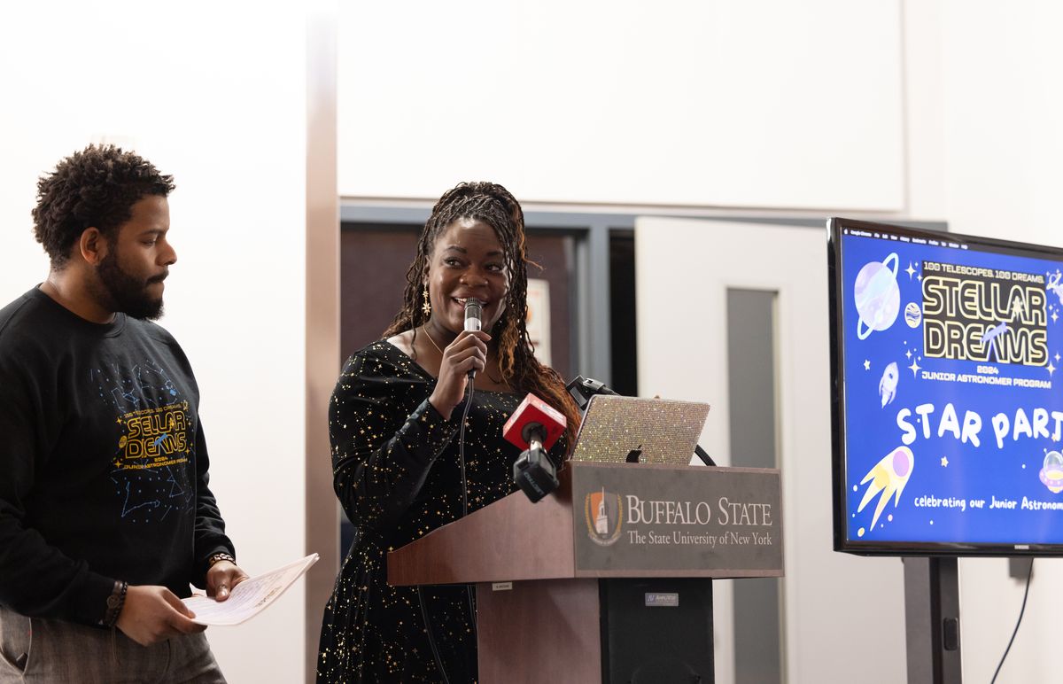 Dr. Raven Baxter speaks into a microphone at the Buffalo State podium as Dr. Ronald Gamble holds the program beside her