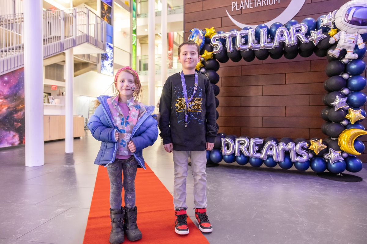 Two young guests, one in a Stellar Dreams sweatshirt and lanyard, stand on the red carpet in front of the balloon arch