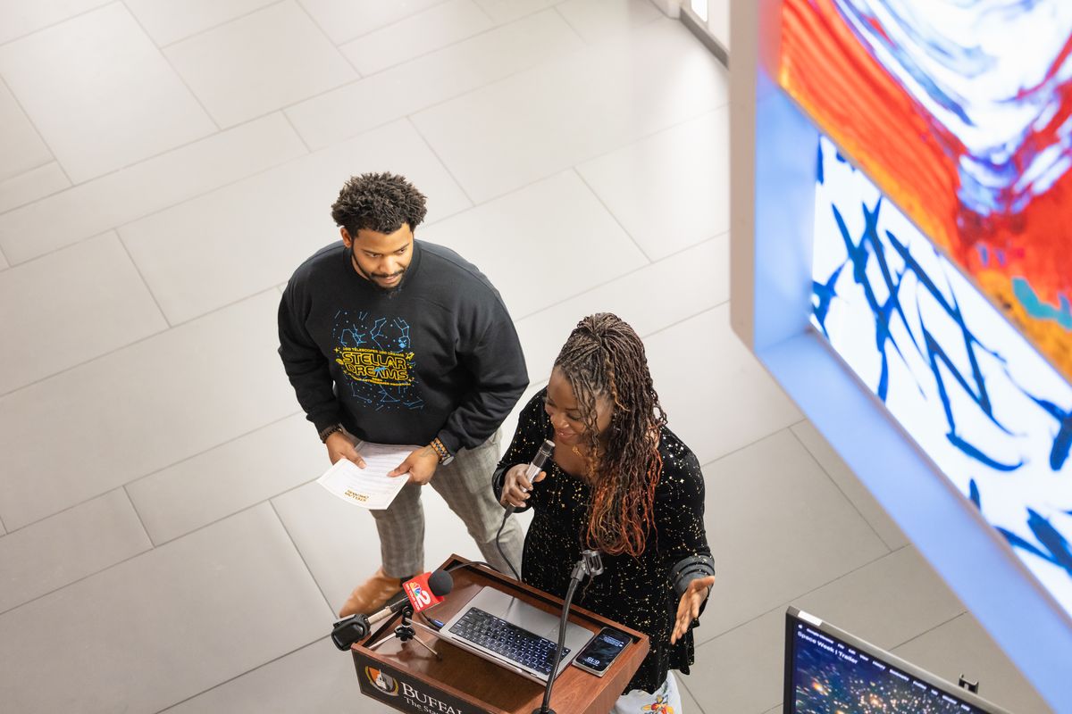 Overhead view of Dr. Raven Baxter speaking at the podium, next to a WGRZ Channel 2 microphone, as Dr. Ronald Gamble holds the program