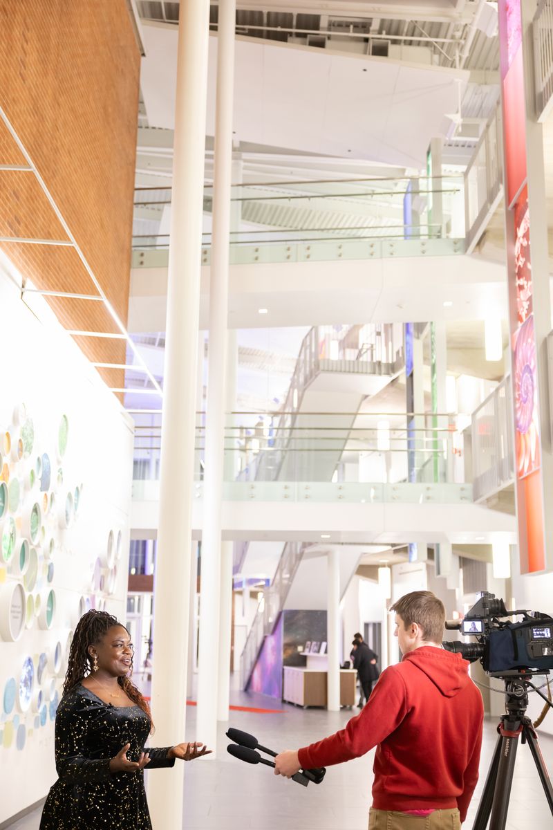 Tall view of the atrium as Dr. Raven Baxter speaks with a WGRZ Channel 2 reporter beside a camera on a tripod, under open balconies