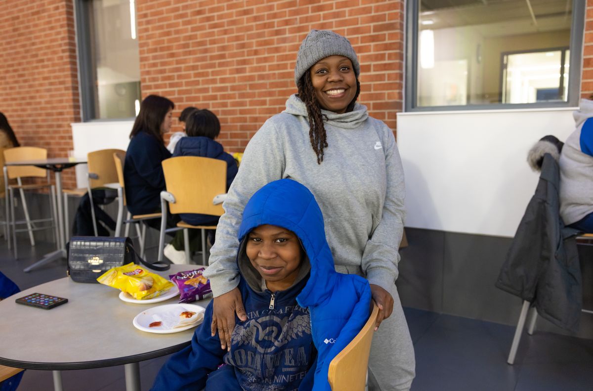 A smiling woman in a gray hoodie stands behind a seated young guest in a blue hooded jacket at a table with snacks