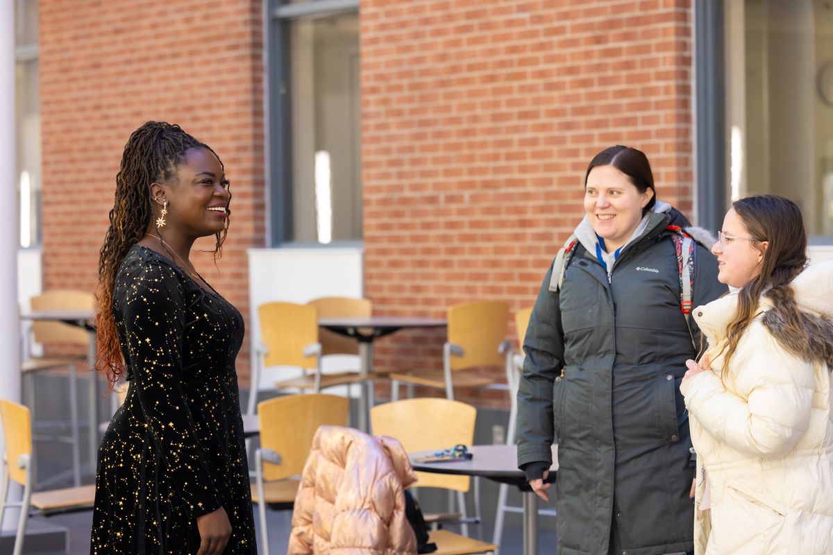 Dr. Raven Baxter, in a black dress with gold stars, smiles as she greets two guests in winter coats in the atrium