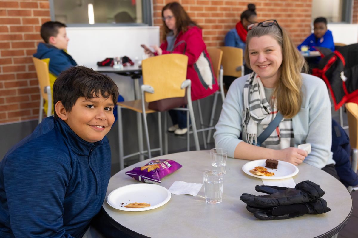A young guest and an adult smile at a table with pizza, a brownie and cups of water before the program