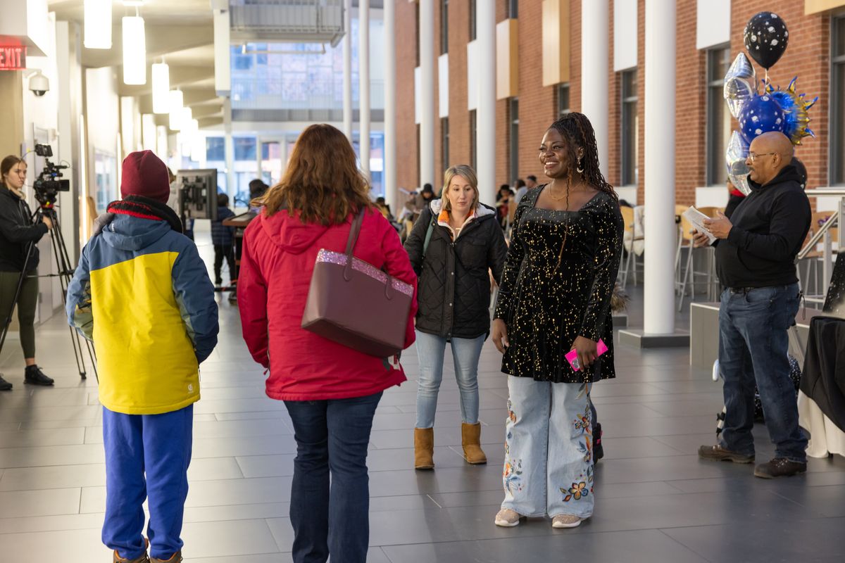 Dr. Raven Baxter smiles at arriving families in the atrium beside a bunch of space balloons, with a video camera at left